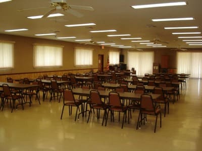 Spacious community hall with numerous tables and chairs arranged neatly, featuring large windows for natural light. Ideal for gatherings and events.