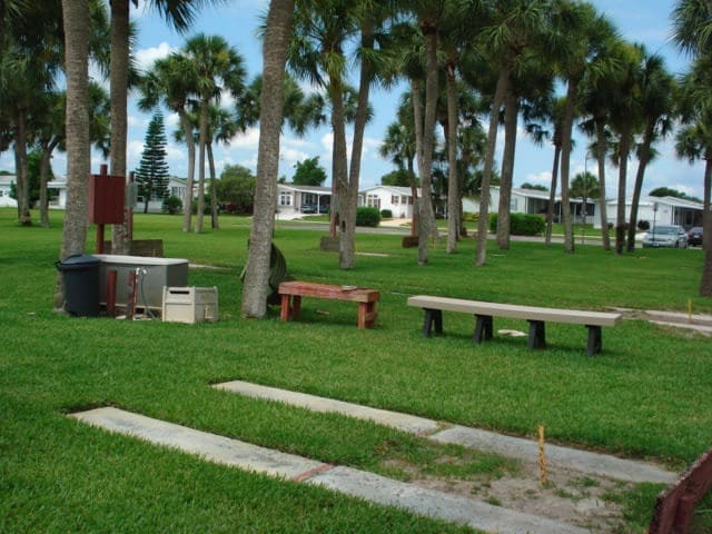 Seating area in a grassy park surrounded by palm trees, featuring two benches and a small trash bin. Mobile homes are in the background.