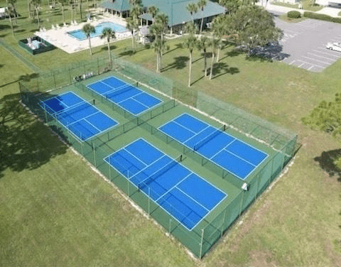 Aerial view of six blue tennis courts surrounded by green grass, with a swimming pool and palm trees in the background. Ideal for recreational activities.