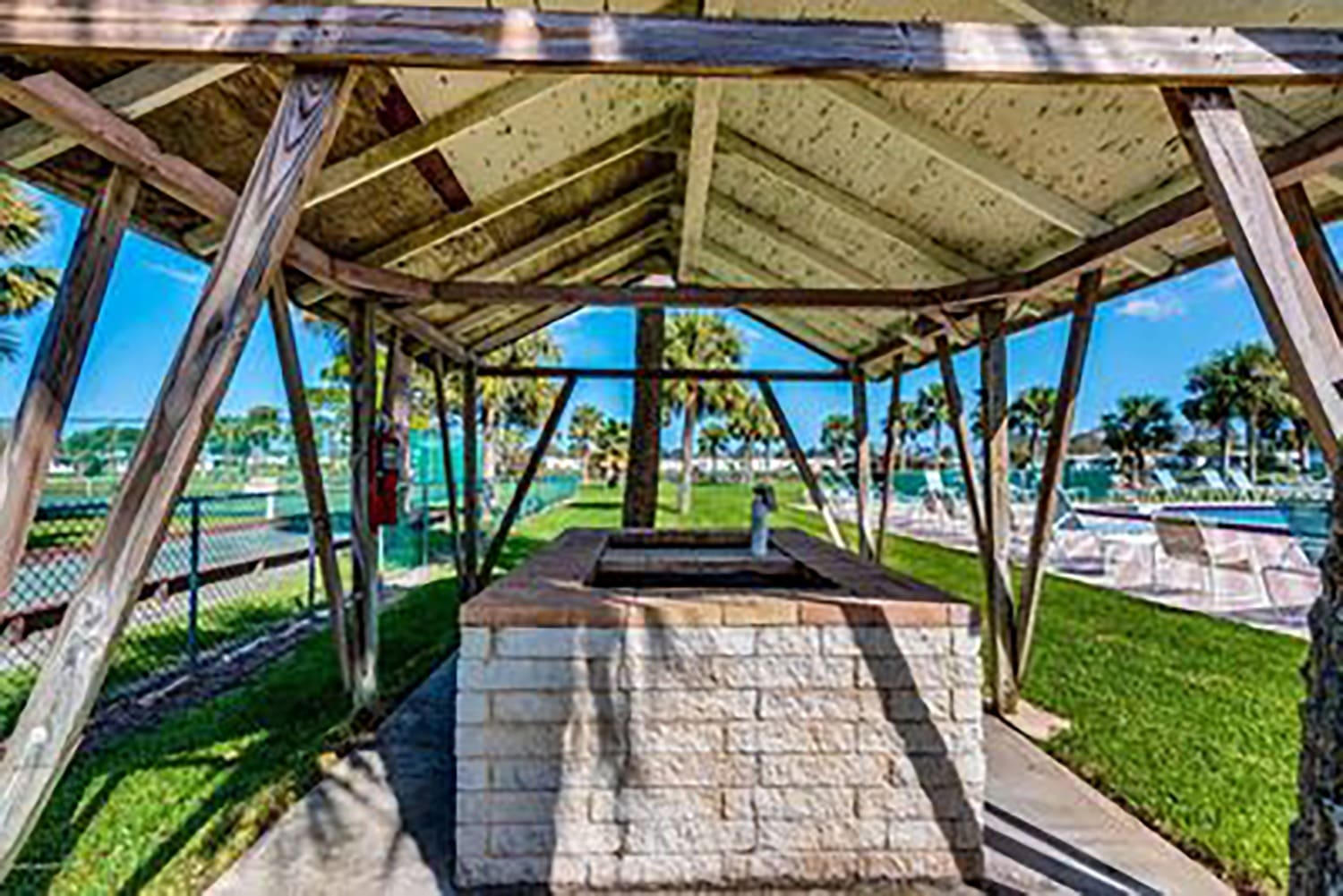 Open-air shelter featuring a stone BBQ pit with a metal grill. Palm trees and lounging areas are visible in the background, highlighting a recreational space.