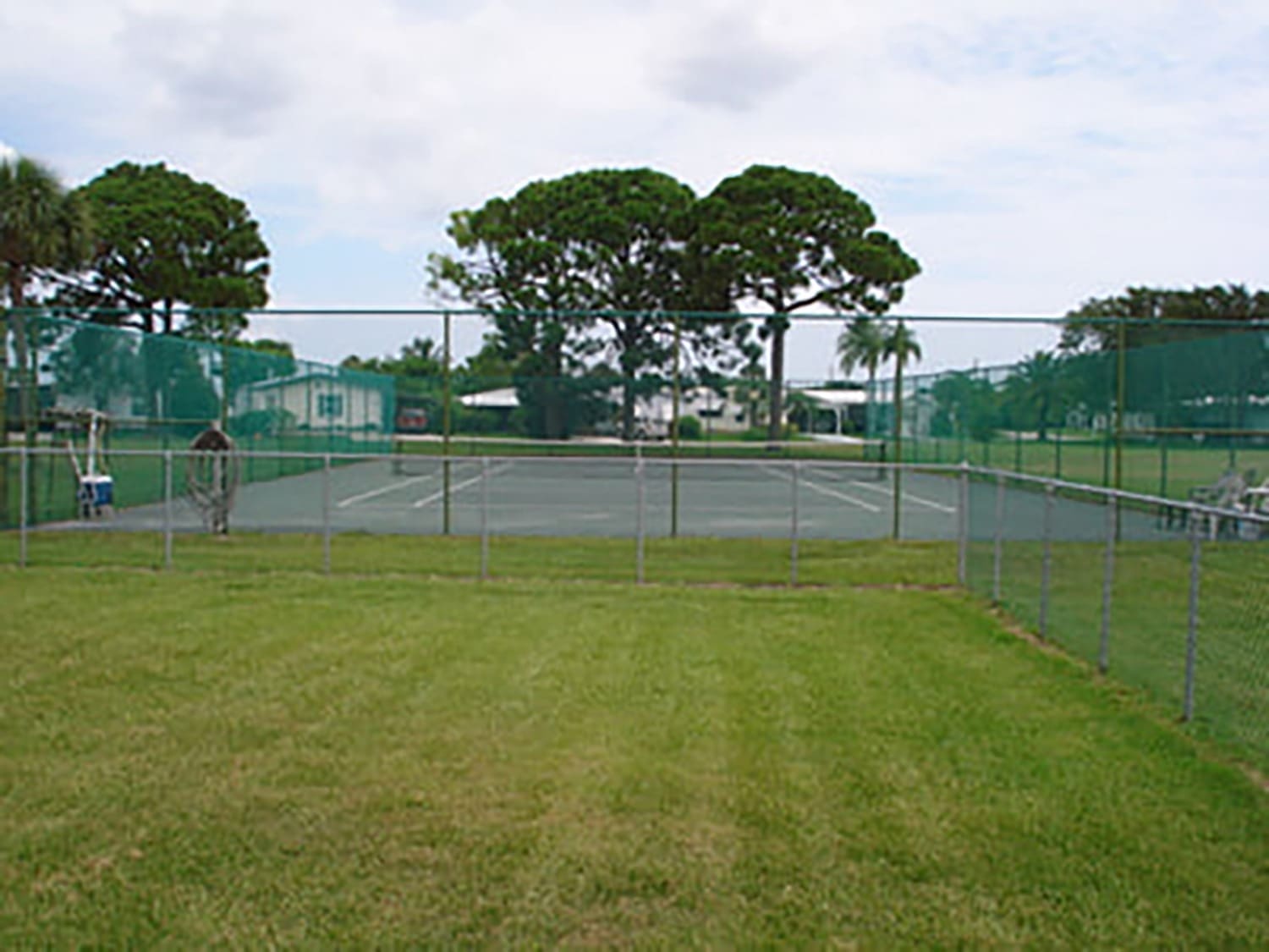 Tennis court enclosed by a fence, with a person walking nearby and lush green grass surrounding the area. Ideal for recreational activities.