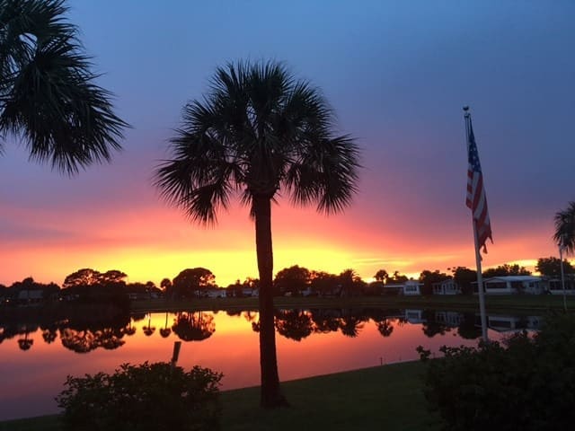 Vibrant sunset reflecting on a calm lake, framed by palm trees and an American flag, showcasing the tranquility of evening in a coastal community.